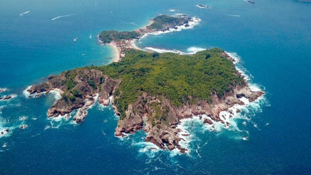 Drone view of Isla Ixtapa off the coast of Zihuatanejo, Mexico, showing lush green hills, turquoise Pacific waters, and sandy beaches.