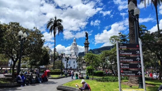 Plaza Grande in Quito, Ecuador, with the Independence Monument, colonial buildings, and directional signs pointing to major attractions.